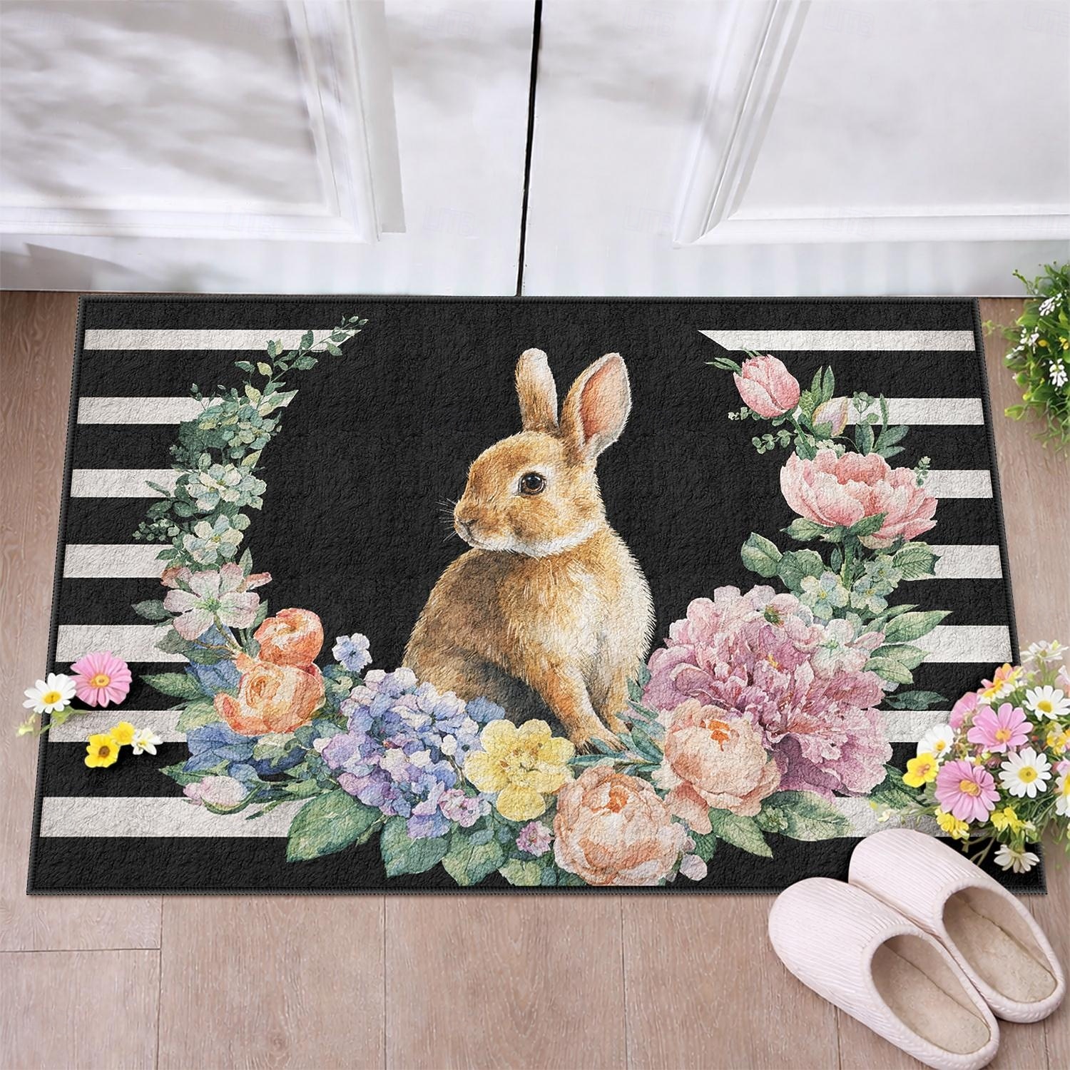 Felpudo de Pascua con forma de conejito rodeado de una corona floral floreciente sobre rayas blancas y negras, perfecto para dar la bienvenida a la primavera. 2026 - $16.99 –P2